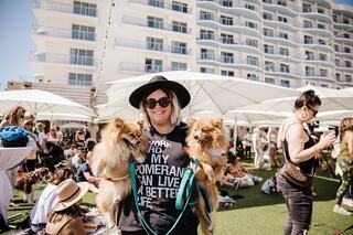 A woman in a black hat holds two Pomeranians at a lively outdoor event with people and umbrellas in the background.