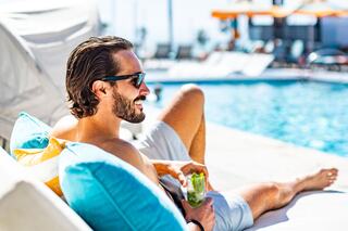 A man relaxes by the pool in shades, enjoying a drink while lounging on colorful cushions, surrounded by a sunny, vibrant atmosphere.