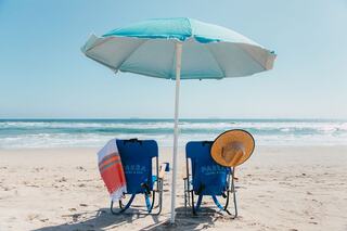 Two beach chairs under a vibrant umbrella, with a towel draped over one and a sun hat resting nearby, facing the ocean waves.