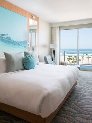 A modern hotel room with a queen bed, ocean view, stylish decor, and large windows showcasing palm trees and a beachscape.