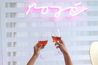 Two hands raise glasses of rosé in front of a glowing pink neon sign that says "rosé," creating a celebratory atmosphere.