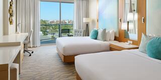 A modern hotel room features two beds, a sleek desk, and large windows with a view of palm trees and clear blue skies.