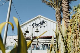 A building sign reads "Pacific City," with palm trees and outdoor seating visible, creating a vibrant, sunny atmosphere.