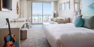 Bright, modern hotel room with a bed, desk, guitar, and stunning ocean view through large windows, enhanced by soft, beachy decor.