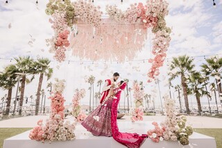 A couple in traditional attire embraces under a floral archway adorned with pink flowers, set against a backdrop of palm trees and a sunny sky.