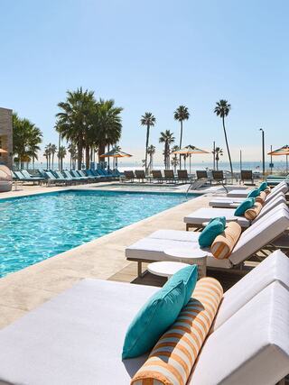 A bright pool area with lounge chairs, tropical palm trees, and blue umbrellas, offering a relaxing vibe near the ocean.
