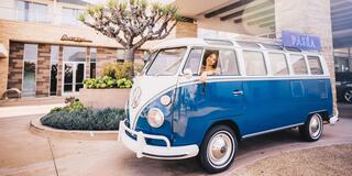 A vintage blue VW bus stands outside a modern building, with a woman leaning out the window, surrounded by lush greenery.