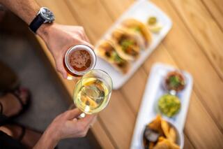 A hand holds a glass of white wine while another holds a pint of beer, accompanied by tacos and chips on a wooden table.
