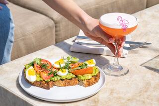 A hand holds a cocktail beside a plate of avocado toast topped with tomatoes, eggs, and greens, set on a sunlit table.