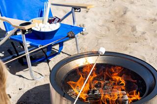 A beach scene featuring a fire pit, marshmallows roasting on sticks, and a nearby chair with snacks, all set in warm sunlight.