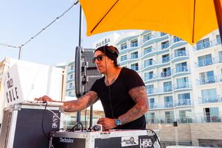 A tattooed DJ spins music under a bright yellow umbrella, with modern buildings in the background, creating a lively atmosphere.