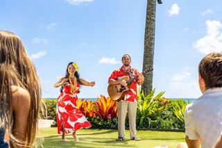 A performer plays guitar while a dancer in a floral dress captivates an audience amidst tropical scenery and bright blue skies.