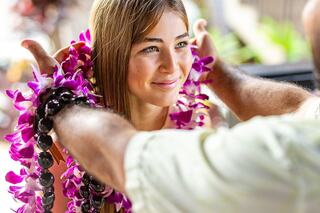 A young woman smiles while receiving a floral lei from someone, embodying a warm, welcoming cultural moment.