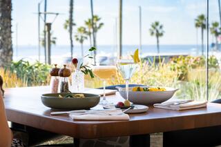 A sunlit table overlooks the ocean, adorned with colorful cocktails and fresh dishes, framed by palm trees and a vibrant backdrop.