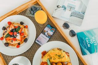 A cozy breakfast spread features pancakes with berries, an omelet, fresh juice, a smartphone, sunglasses, and books on calmness and decor.