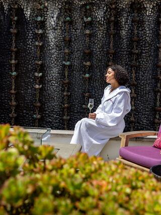 A woman in a white robe enjoys a glass of champagne, relaxed by a textured stone wall and vibrant greenery, exuding tranquility and leisure.
