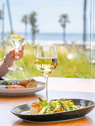 A stylish dining scene features a glass of white wine, a plate of vibrant asparagus and culinary delicacies, with a beach view in the background.
