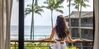 A woman gazes at the ocean, surrounded by palm trees and a tropical resort, exuding a sense of peace and relaxation.