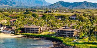 A scenic coastal view featuring beachfront accommodations surrounded by palm trees, lush greenery, and majestic mountains in the background.
