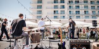 A lively outdoor concert features a band performing for a captivated crowd, with an array of tents and a hotel in the background.