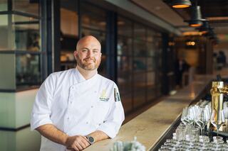 A smiling chef in a white uniform stands confidently at a bar, surrounded by glassware, exuding professionalism and warmth.