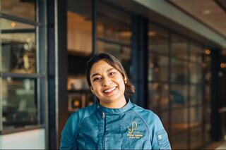 A smiling chef in a professional culinary uniform stands in a modern kitchen, exuding warmth and passion for her craft.