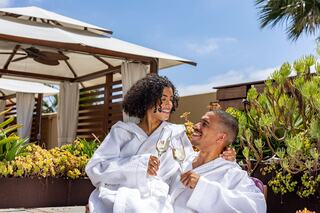 A joyful couple in white bathrobes shares a toast with champagne, surrounded by lush greenery and a sunny, relaxing atmosphere.