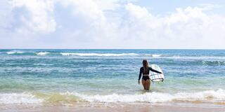 A person in a black swimsuit walks towards the ocean, carrying a surfboard under a bright sky with fluffy clouds. Waves gently lap at the shore.