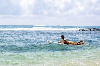 A person paddles on a surfboard in clear blue waters, surrounded by gentle waves and a bright, cloudy sky.