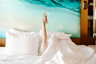 A hand holding a cocktail glass emerges from a cozy bed, surrounded by fluffy pillows and a serene, aqua-themed backdrop.