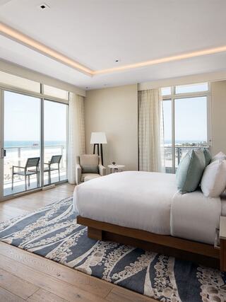 A modern, airy bedroom features a large bed, soft colors, a cozy seating area, and a beautiful view of the ocean through sliding glass doors.