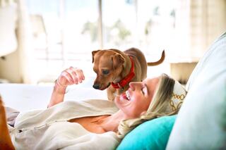 A smiling woman relaxes on a bed while a playful dog approaches her, creating a joyful and affectionate moment.