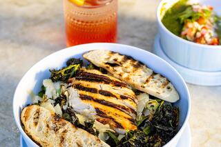 A bowl of grilled chicken over vegetables with greens, accompanied by toasted bread and a refreshing drink nearby.