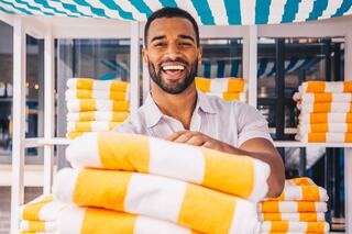 A smiling man stands behind a stack of neatly arranged orange and white towels, dressed casually in a light shirt, exuding warmth and hospitality.