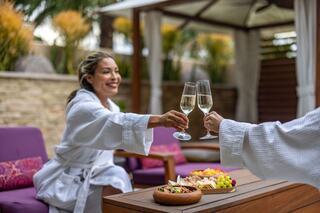 Two women in bathrobes cheerfully clink glasses of champagne while enjoying a relaxing, outdoor gathering with snacks.