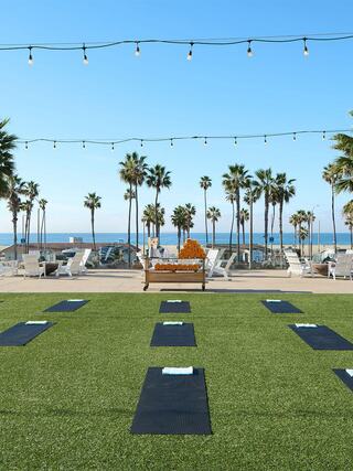 A serene outdoor yoga space features mats on grassy turf, surrounded by palm trees and ocean views, with string lights overhead.