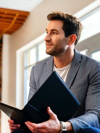A man in a gray blazer sits thoughtfully, holding a laptop and looking off to the side, surrounded by natural light.