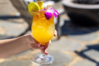 A tropical cocktail garnished with a purple flower and pineapple sits beside colorful flip-flops and a poolside menu, adding a relaxed vibe.