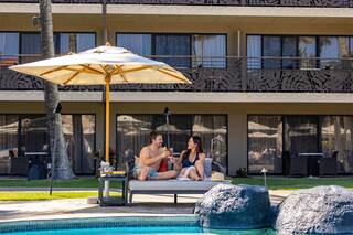 A couple enjoys drinks on a poolside lounge beneath an umbrella, surrounded by lush greenery and a modern resort backdrop.