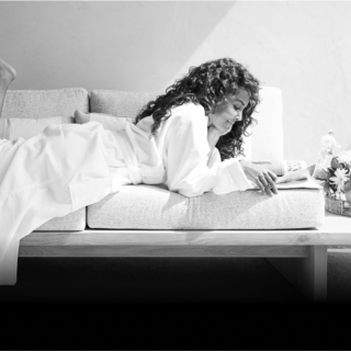 A woman in a robe relaxes on a couch, lost in a book under warm light, with flowers in a basket nearby, exuding calm and tranquility.