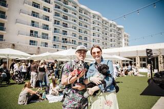 A cheerful couple holds small dogs while surrounded by people enjoying a sunny outdoor event near a modern building.