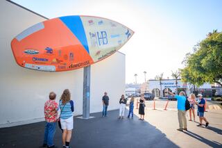A large surfboard installation on a wall attracts a group of onlookers, highlighting surf culture and community engagement.
