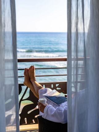 Bare feet rest on a balcony railing, with a book in one hand and a coffee cup in the other, overlooking a serene ocean view.