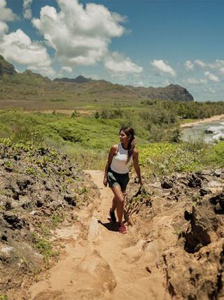 A woman strolls along a sandy trail, surrounded by lush greenery and rocky terrain, with mountains and a coastline in the background.