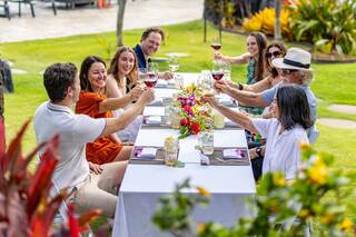 A festive gathering outdoors with people toasting over a long table adorned with flowers. Laughter and joy fill the atmosphere.