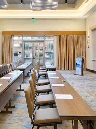 A spacious meeting room features several rows of tables and chairs, bright windows, and warm lighting, creating a professional atmosphere.