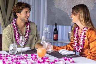 A couple enjoys an intimate dining experience adorned with tropical flowers, sharing smiles and conversation, surrounded by elegant table settings.