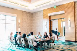 A group meets around a table in a modern conference room, while someone enters carrying a surfboard.