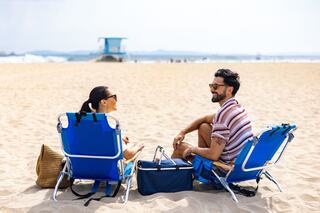 A couple enjoys a sunny day at the beach, sitting in lounge chairs, smiling, and chatting, with a lifeguard tower and ocean in the background.