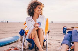 A woman with curly hair smiles while holding a drink, relaxed in a beach chair, enjoying a sunny day with friends near the ocean.
